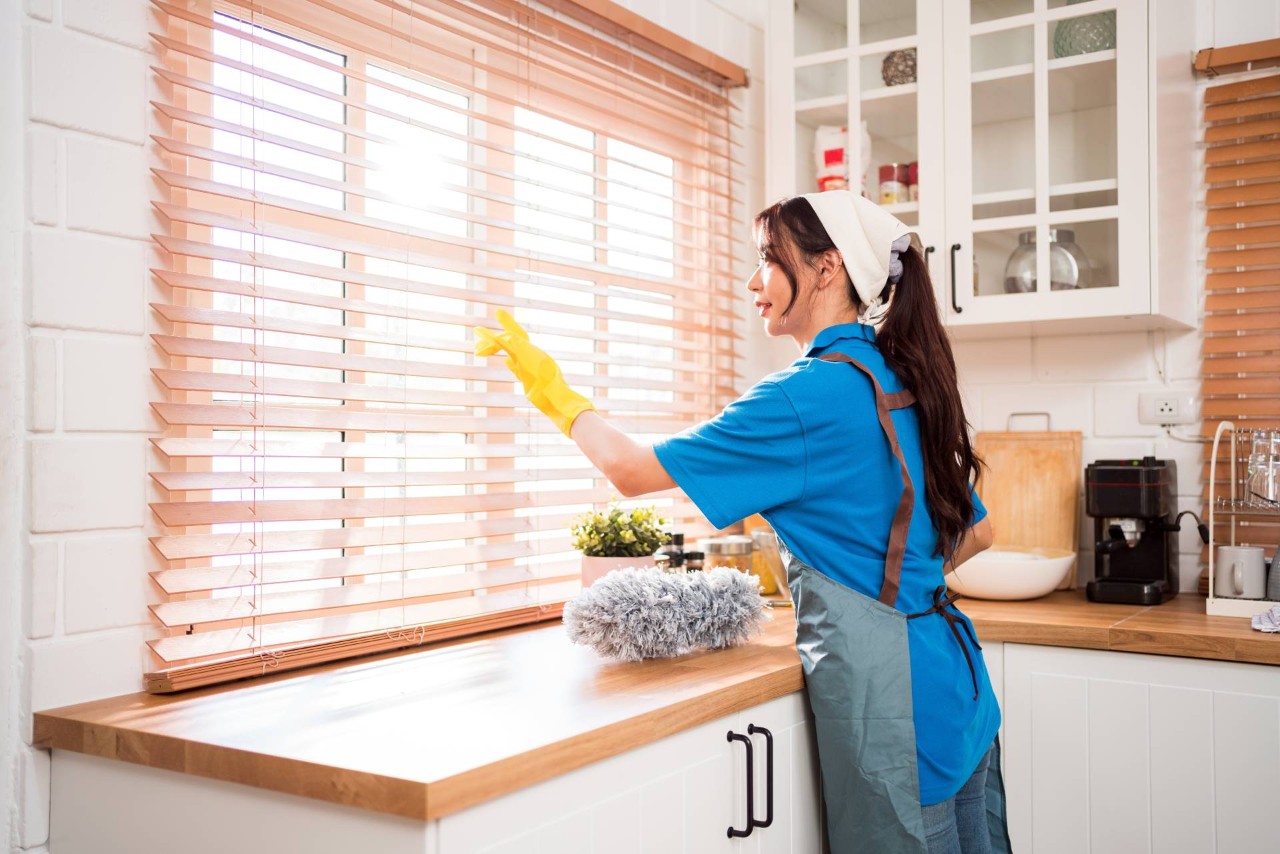 Woman wearing yellow gloves cleaning her wood window blinds with a feather duster in a kitchen at Custom Window Decor of Jupiter near Jupiter, FL